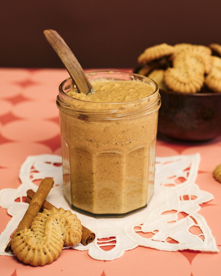 Lasipurkki täynnä kanelista piparkakkumaista kaneliässätahnaa, lusikka purkissa; edessä kanelitankoja ja kaneliässiä vaalealla pitsiliinalla, taustalla kulho täynnä pikkuleipiä vaaleanpunaisella kuviopöydällä.
