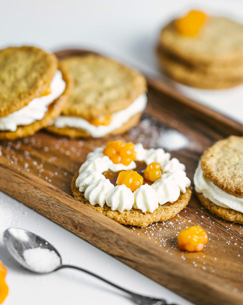 Kaurainen kerrosleivos Lähikuva puisella tarjoilualustalla olevasta pyöreästä kauraisesta kerrosleivoksesta tai pikkuleivästä. Kaksi kauraista keksiä tai pohjaa on täytetty runsaalla kermavaahdolla ja kirkkaan oranssilla lakkahillolla. Yksi leivos on koristeltu pursotetulla kermavaahtokehällä, keskellä lakkahilloa ja päällä tuoreita lakkoja. Kuva on tyylitelty, ja valkoinen tausta korostaa makeisten ruskeita ja oransseja sävyjä.