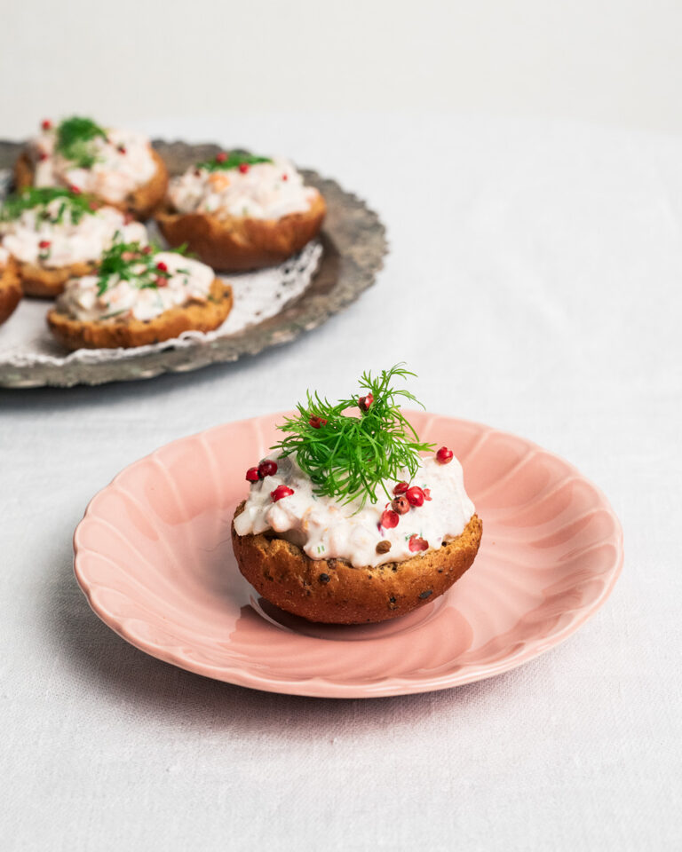 Saaristolaiskorppu, jonka päällä on kermainen kylmäsavulohitäyte, roseepippureita ja tuoretta tilliä vaaleanpunaisella lautasella; taustalla tarjotin täynnä lohikorppuja valoisalla, neutraalilla pöydällä.
