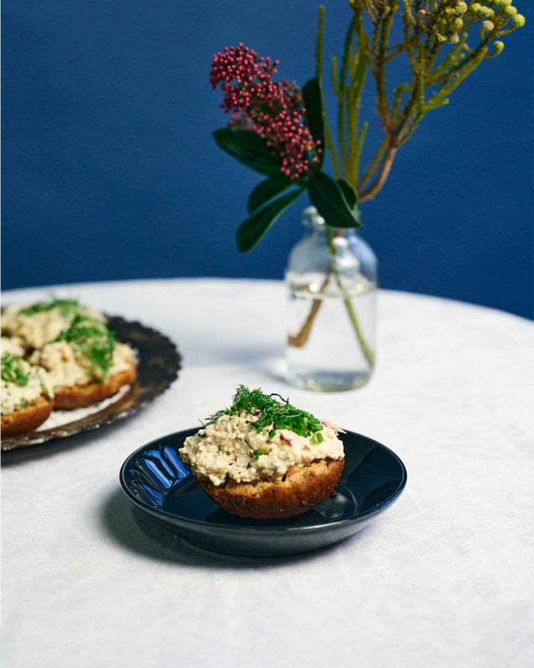 Saaristolaisleivästä tehdyt herkulliset tofuskagenkorput tarjoiluvadilla; korppujen päällä runsas vegaaninen tofutäyte sekä tuoretta tilliä, edessä yksi korppu lautasella ja taustalla maljakko kukkineen sinistä taustaa vasten.