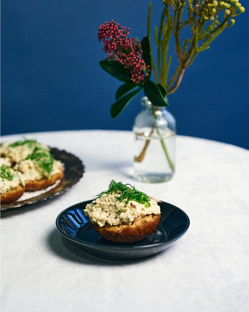 Saaristolaisleivästä tehdyt herkulliset tofuskagenkorput tarjoiluvadilla; korppujen päällä runsas vegaaninen tofutäyte sekä tuoretta tilliä, edessä yksi korppu lautasella ja taustalla maljakko kukkineen sinistä taustaa vasten.