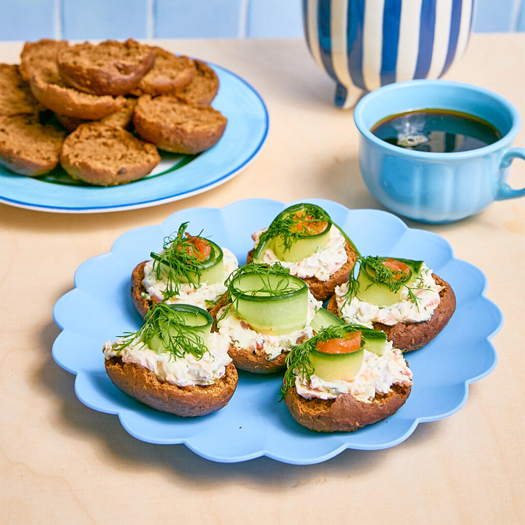 K. Leivon Saaristolaiskorput lohitäytteellä, koristeltu kurkulla ja tillillä, aseteltuina siniselle lautaselle, kahvikuppi taustalla.