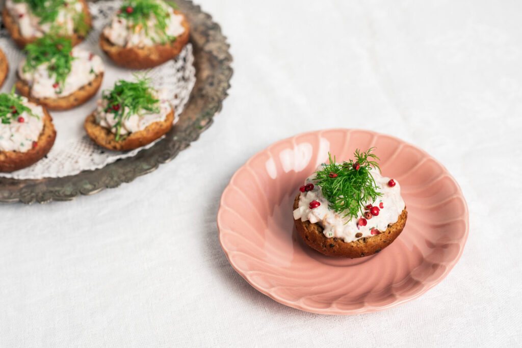 K. Leivon saaristolaiskorput savulohitäytteellä, koristeltu tillillä ja rosepippurilla, tarjoiltuna vaaleanpunaisella lautasella osana juhlapöytää.