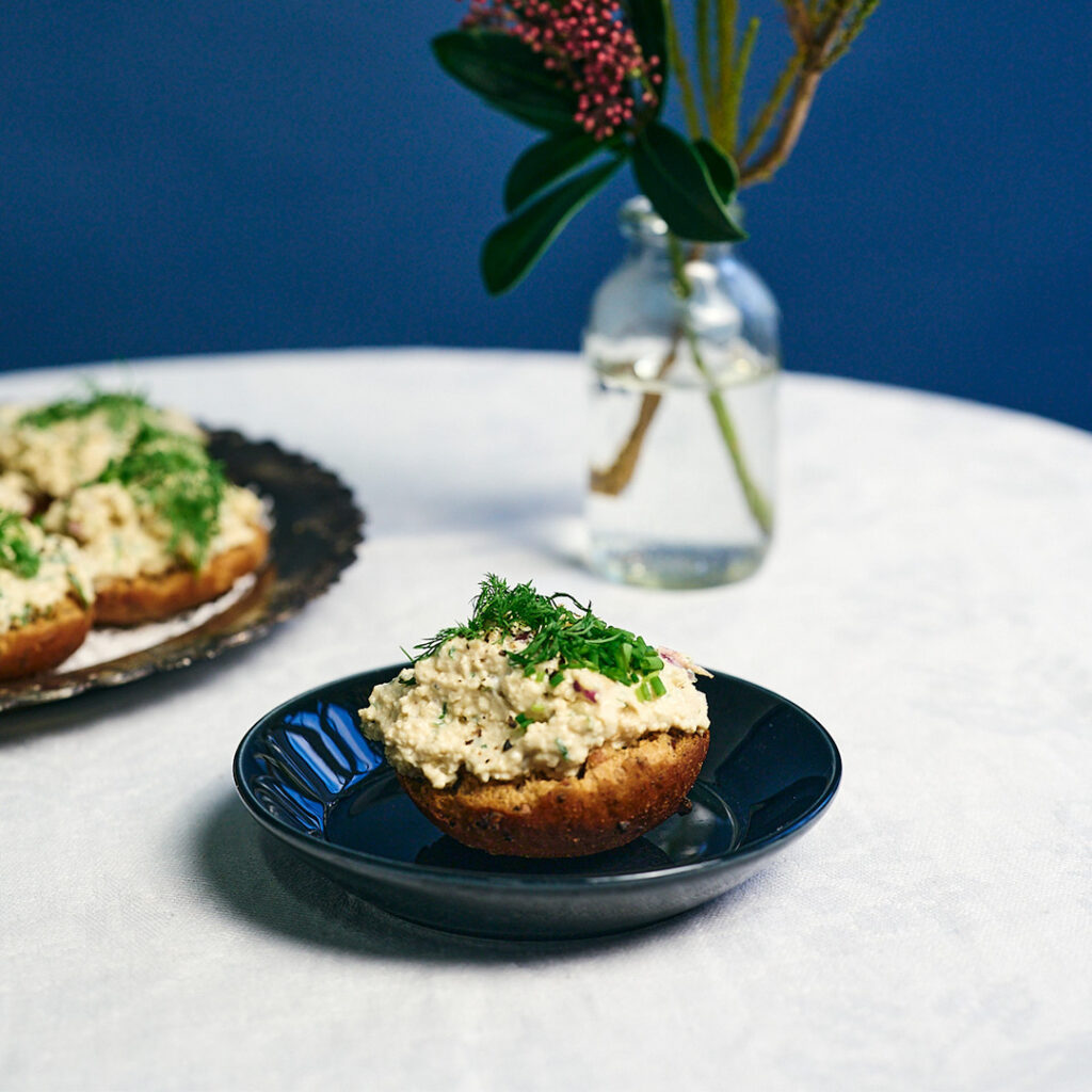 K. Leivon saaristolaiskorppu vegaanisella tofuskagenilla, koristeltu tillillä ja ruohosipulilla, tarjoiltuna tummalla lautasella tyylikkäässä kattauksessa.